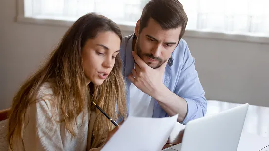 A man and a woman sitting together at a desk, reviewing documents and looking at a laptop in a home office setting.