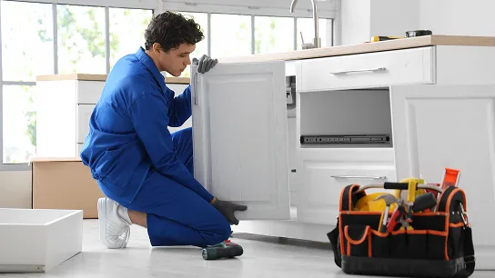 A person in blue coveralls is kneeling and installing a white cabinet door in what appears to be a kitchen. A toolbox sits on the floor nearby.